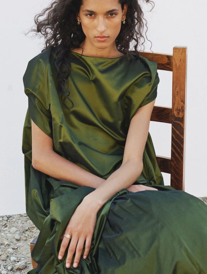 Model seated on a wooden chair wearing a lustrous green satin dress against a plain white background