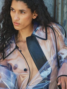 Model with long dark curly hair wearing a translucent vinyl raincoat with black trim against an industrial metal background.