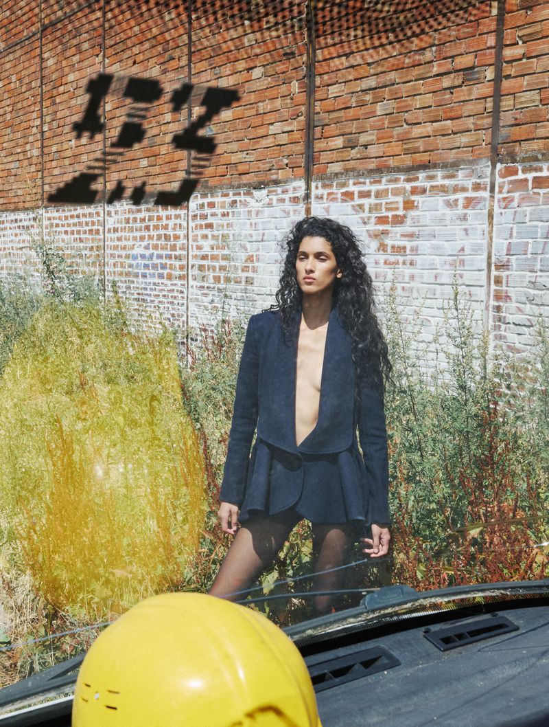 Model with long dark curly hair wearing a blue jacket standing against a brick wall with overgrown weeds.