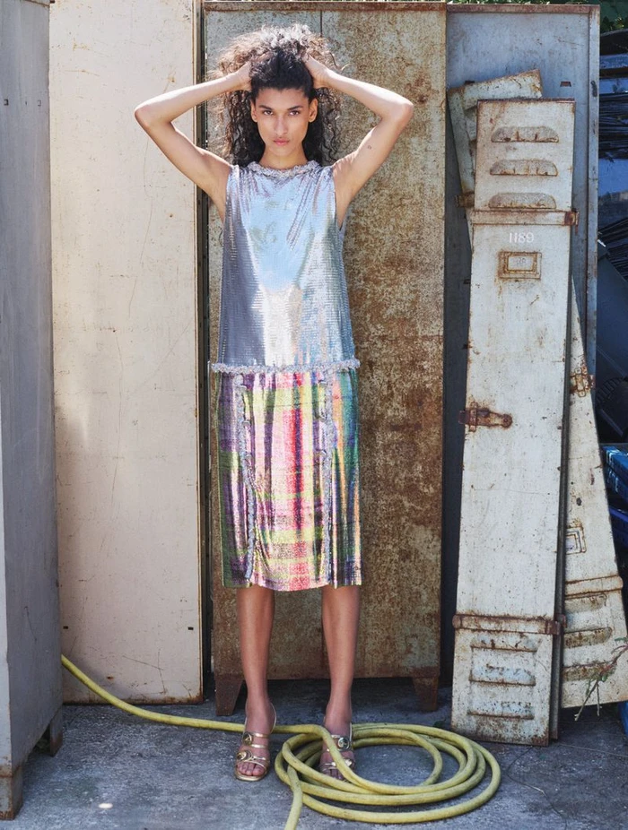Model with dark curly hair posing in a metallic sleeveless dress standing against rusty metal lockers with a yellow hose.