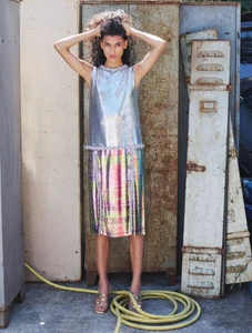 Model with dark curly hair posing in a metallic sleeveless dress standing against rusty metal lockers with a yellow hose.