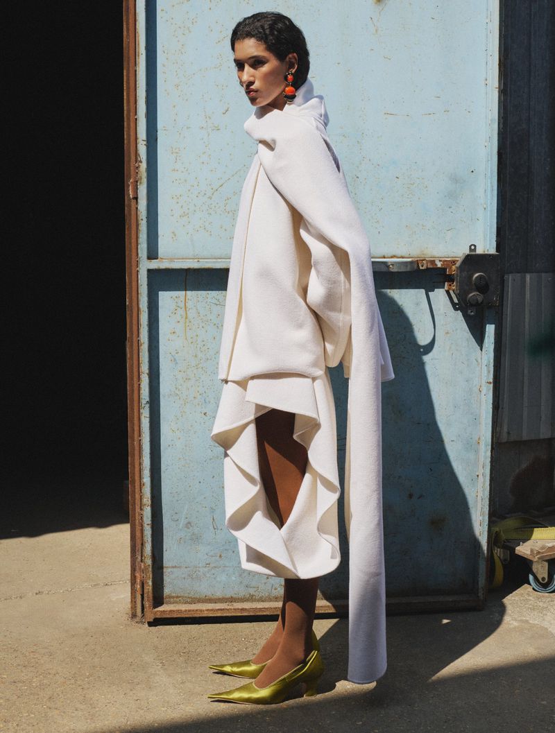 Model wearing a structured white coat and yellow heels standing before a weathered blue door