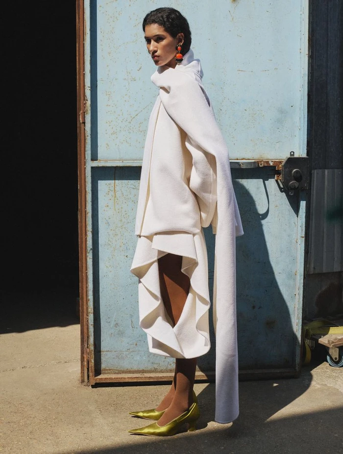 Model wearing a structured white coat and yellow heels standing before a weathered blue door