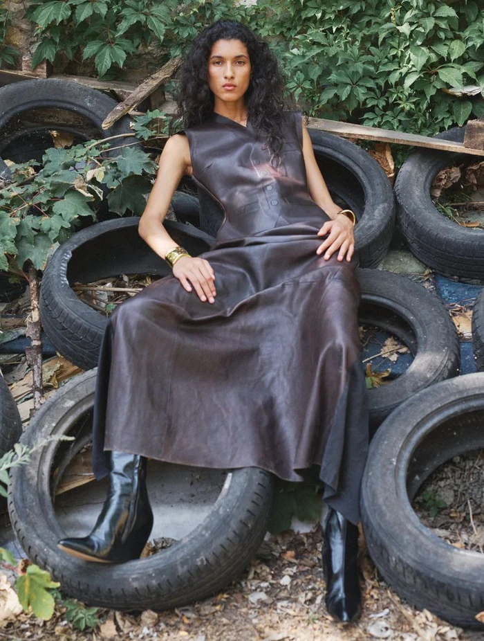 Model seated among stacked black tires wearing a dark leather sleeveless dress and boots for CR Fashion Book