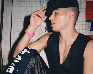 Young man wearing a black cap, black tank top, and printed trousers with a pink wristband, sitting indoors.