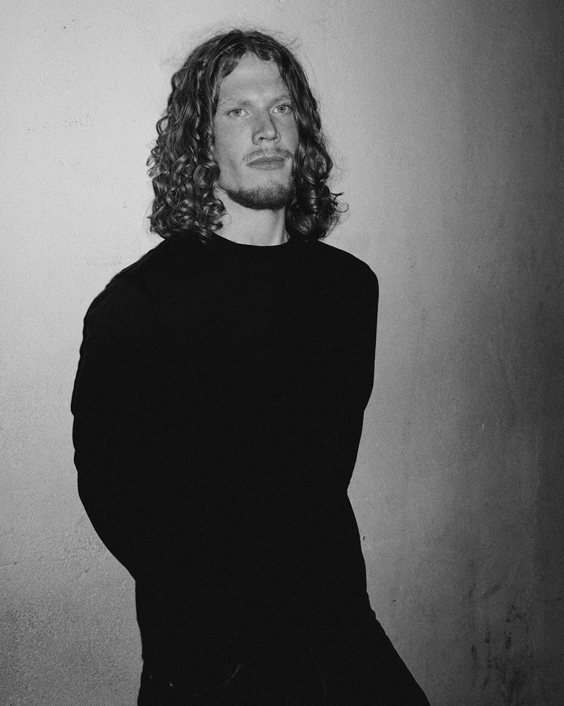 Portrait of a young man with long curly hair and beard wearing a dark long-sleeve shirt standing against a wall