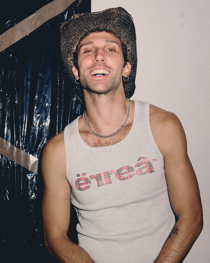 Smiling man wearing a textured cowboy hat and a white tank top with red graphic text, photographed against a white wall.
