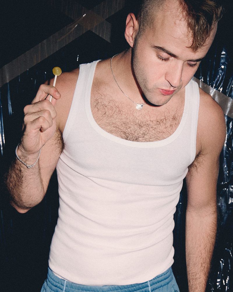 Man wearing a white tank top holding a lollipop against a shiny crinkled metallic background