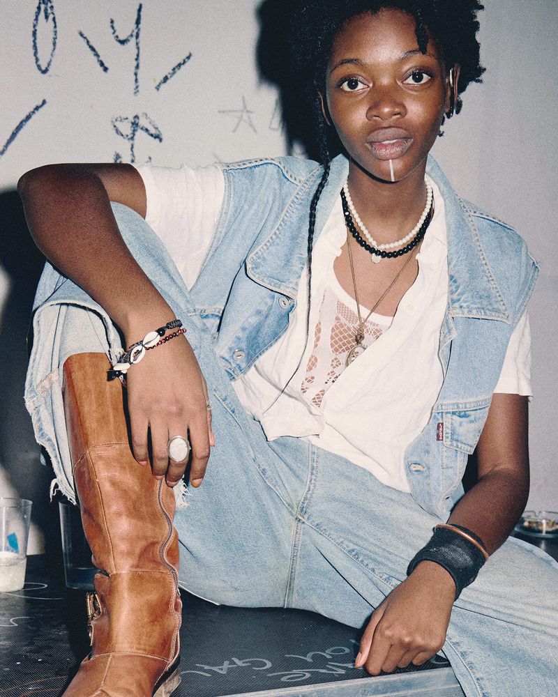 Portrait of a Black woman wearing a denim vest, light jeans, layered necklaces, and tall brown leather boots against a white wall.