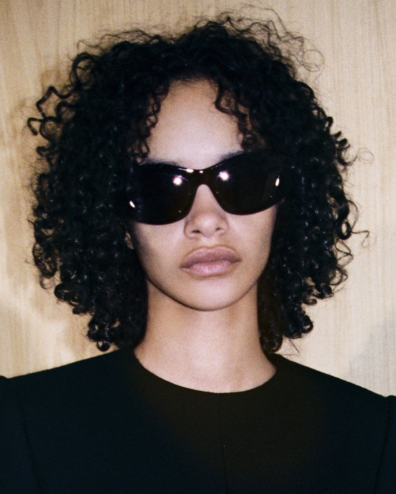Close-up portrait of a young adult woman with curly black hair wearing sunglasses and a black shirt, against a light wood background.