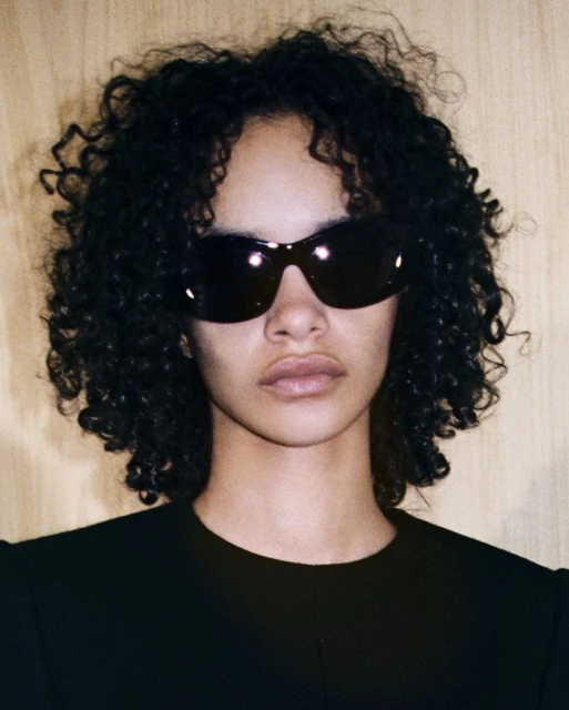 Close-up portrait of a young adult woman with curly black hair wearing sunglasses and a black shirt, against a light wood background.