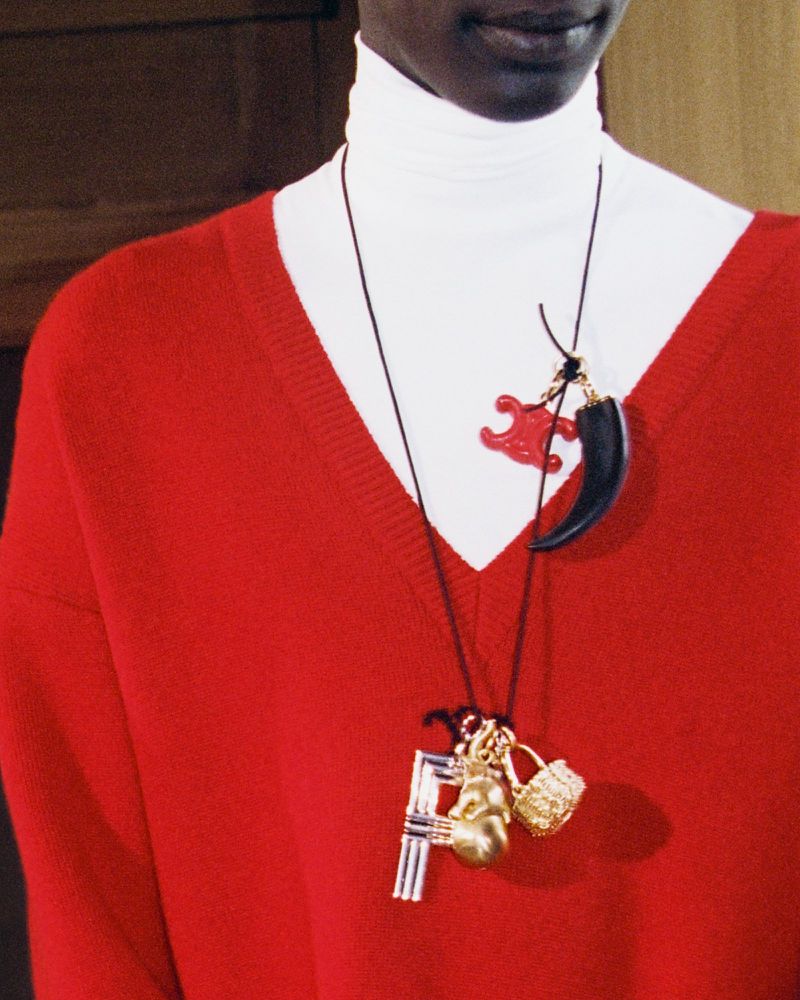 A dark-skinned model wears a red sweater and white turtleneck, accessorized with a necklace featuring multiple charms.