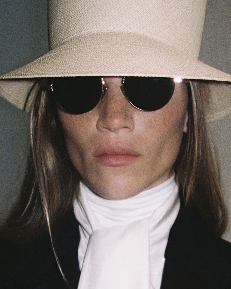 Woman wearing a straw hat and black sunglasses, gazing intently at the camera, dressed in a black jacket and white scarf.