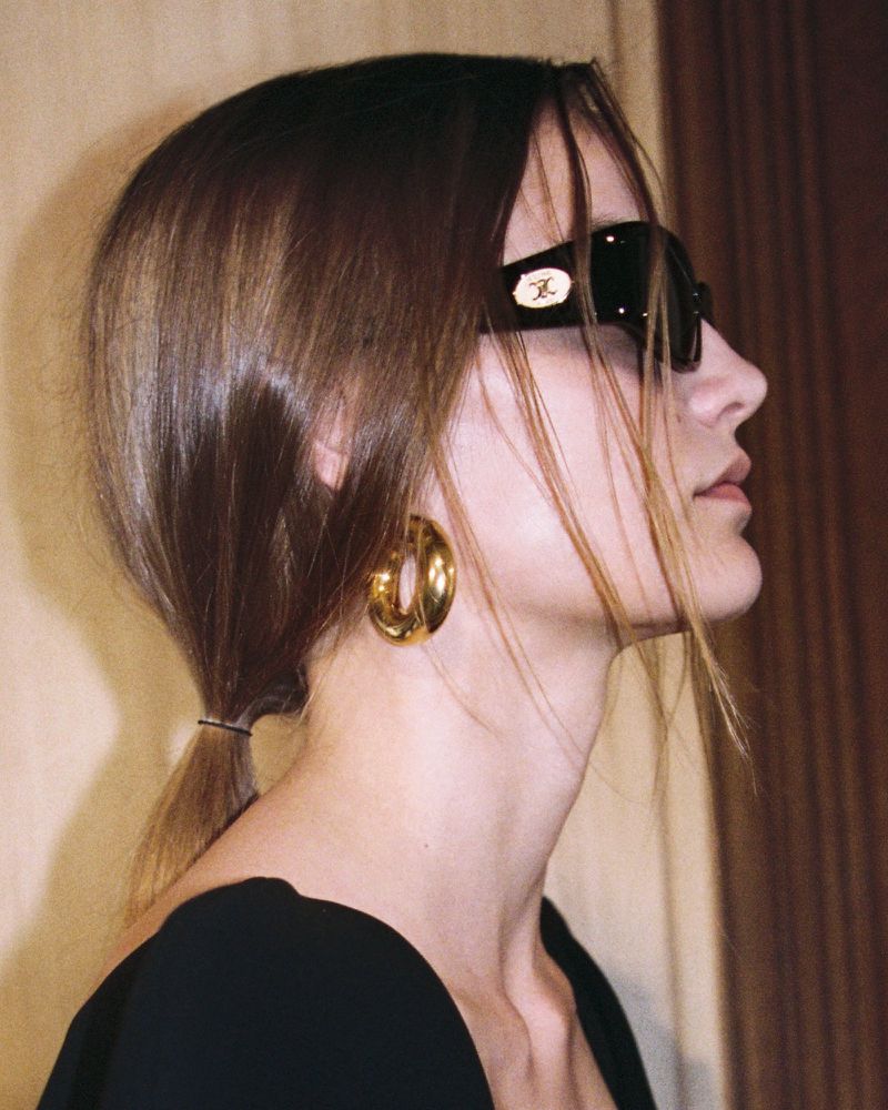 Profile shot of a woman wearing black sunglasses and large gold hoop earrings. She is wearing a black top indoors.