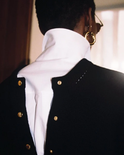 Back view of a Black woman in a black top and white turtleneck, gold hoop earrings, and eyeglasses, standing indoors near a wood wall.