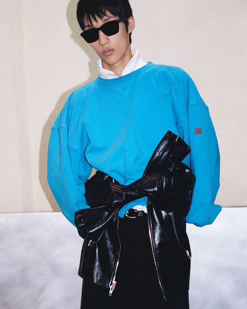 An Asian adult male in sunglasses wears a bright blue sweatshirt with a black leather jacket tied around his waist in a studio setting.