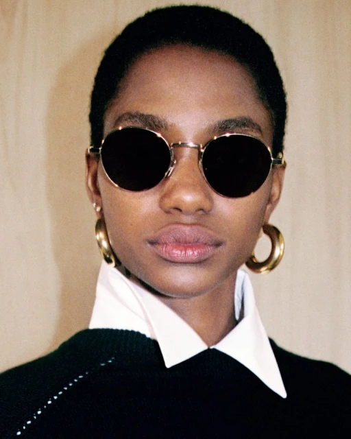 Close-up shot of an African American woman with round sunglasses, gold hoop earrings, and a white collared shirt under a black sweater.