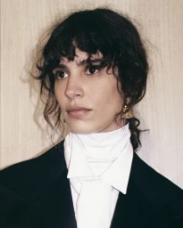 Close-up portrait of a woman with dark wavy hair wearing a white high-neck collar and dark blazer against a light wooden wall