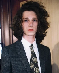 Close-up portrait of a male model with long dark curly hair wearing a dark patterned suit, white shirt, and floral tie backstage at Celine.