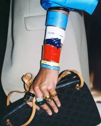 Close-up of a model's arm stacked with colorful branded cuffs and rings, holding a black handbag for Celine