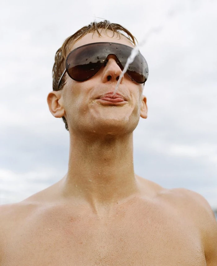 Close-up portrait of a man wearing dark sunglasses with a stream of water splashing near his mouth outdoors.