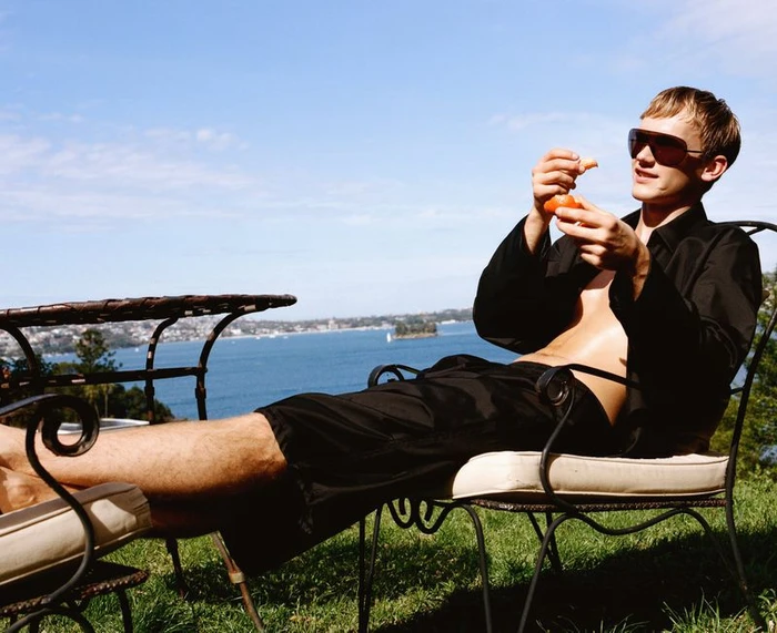 Model wearing sunglasses and black clothing relaxing outdoors by the water for GQ Australia