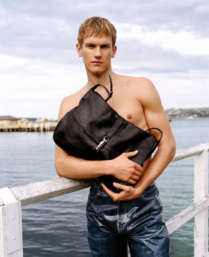 Shirtless male model wearing dark denim jeans leaning on a white railing holding a black leather bag near water