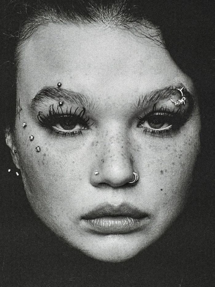 Close-up portrait of a woman with facial piercings, heavy eyelashes, and freckles in monochrome film grain.