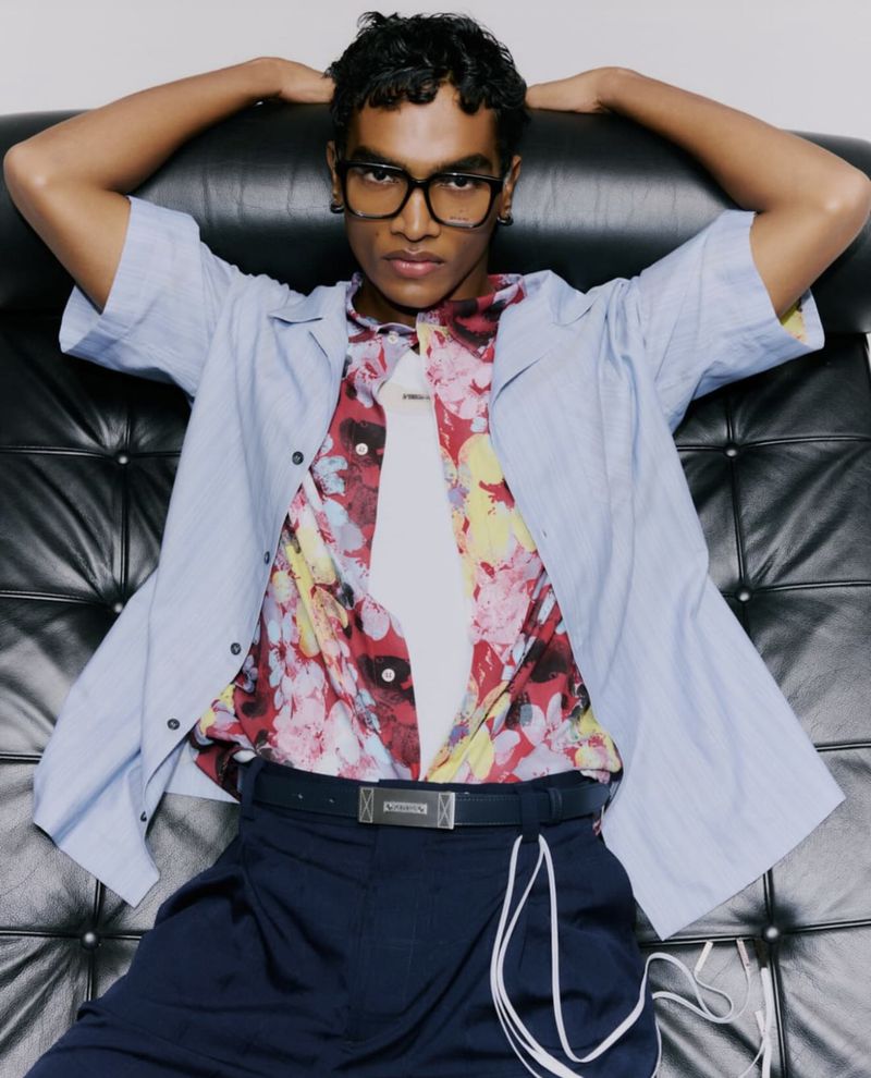 Portrait of a young adult male wearing glasses and a floral shirt, reclining on a black tufted chair in a brightly lit indoor setting.