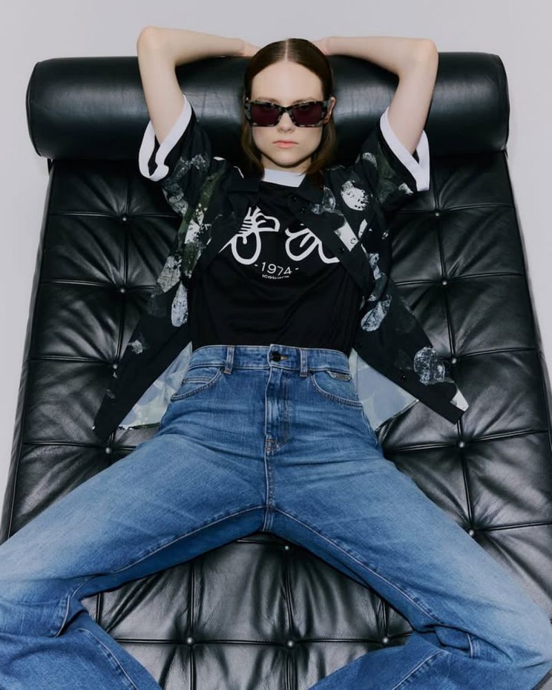 A young adult woman with sunglasses poses on a black leather couch, wearing jeans, a graphic t-shirt and a black-and-white patterned shirt.