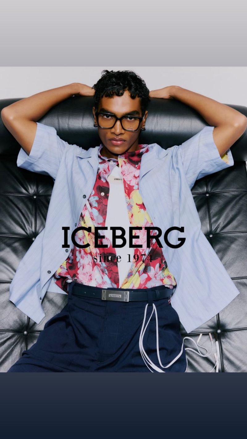 Man poses in a patterned shirt and navy pants on a black textured chair, wearing eyeglasses, with 'ICEBERG since 1974' text overlay.