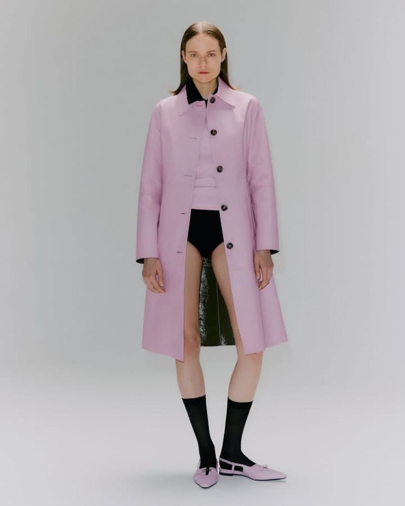 A fair-skinned adult woman poses in a pink trench coat, black top and bottom, and matching pink flats against a pale gray background.