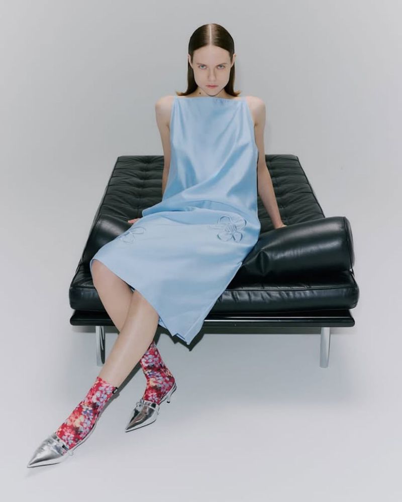 A young woman with straight brown hair is sitting on a black leather couch in a light blue dress with floral socks and silver heels.