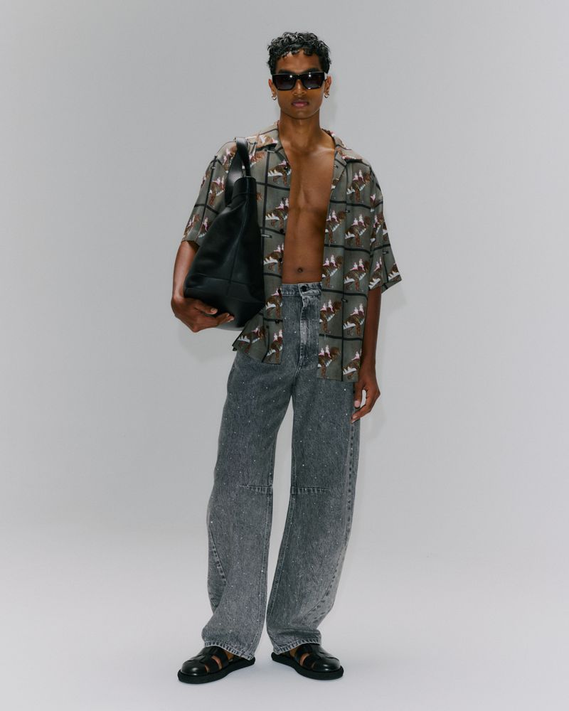 Man in patterned shirt and grey jeans holding a black tote bag, posing in a full shot against a white backdrop, wearing sunglasses.