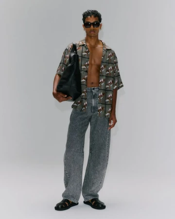 Man in patterned shirt and grey jeans holding a black tote bag, posing in a full shot against a white backdrop, wearing sunglasses.