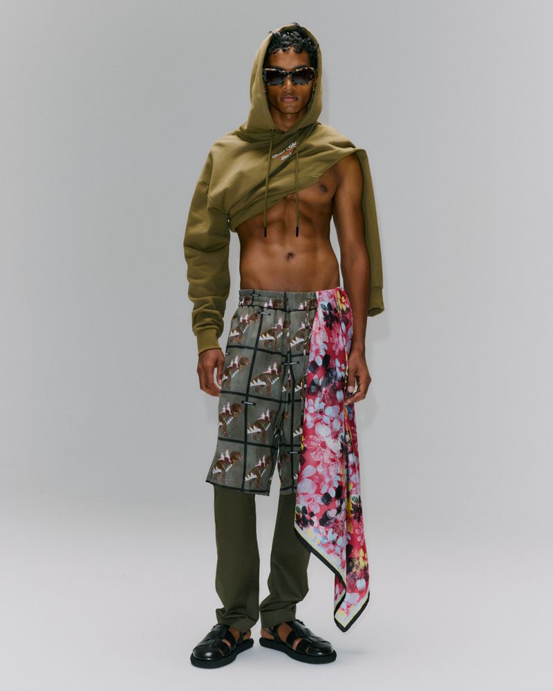 Man in hoodie and patterned shorts poses in a fashion shot against a white background. He wears sunglasses and sandals.