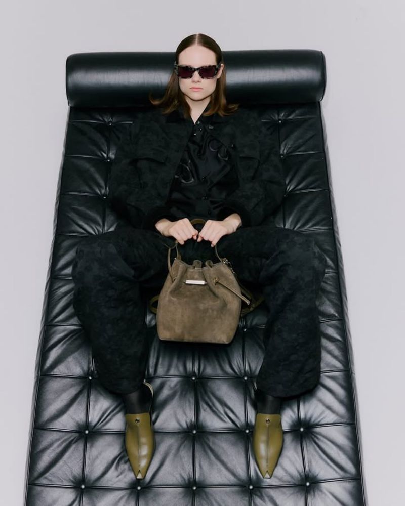 A woman in sunglasses and a patterned black outfit is seated on a black leather bench holding a brown suede bucket bag.