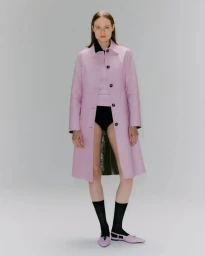 Model wearing a long pink coat with black undergarments and dark socks against a grey studio backdrop