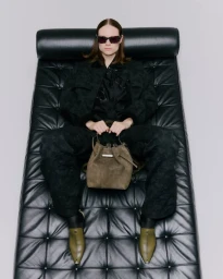 Model wearing dark sunglasses and black apparel seated on a black leather lounge chair holding a bucket bag