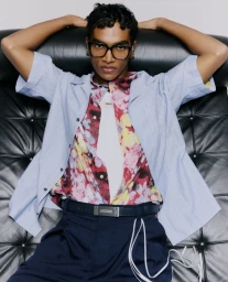 Model wearing black framed glasses, blue button down shirt, and floral shirt lounging on a black leather sofa.
