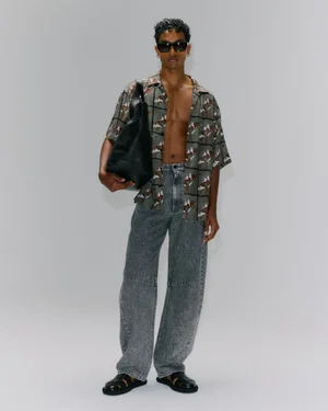 Male model in patterned short-sleeve shirt and grey denim jeans holding a large black leather bag against a white seamless backdrop.