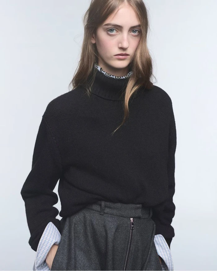 Model wearing a black turtleneck sweater and grey trousers against a plain white studio background.