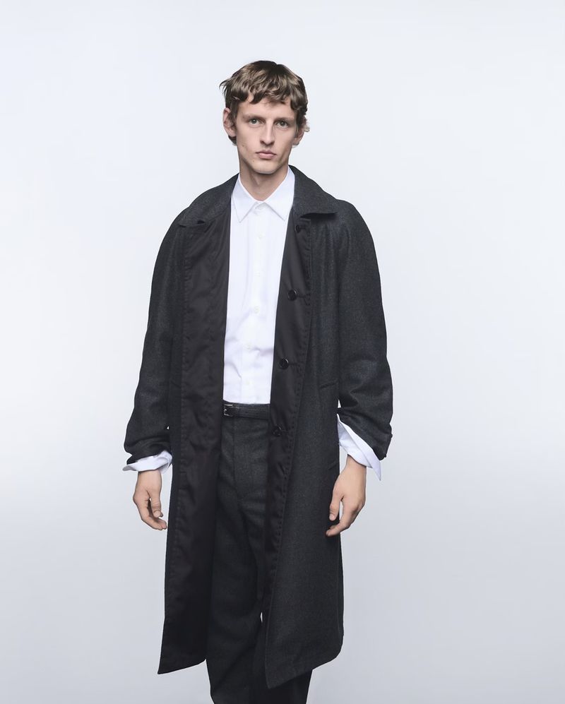 Model wearing a dark coat over a white shirt against a plain white studio background