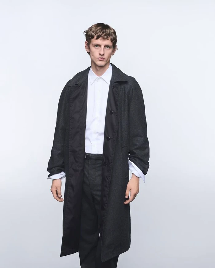 Model wearing a dark coat over a white shirt against a plain white studio background