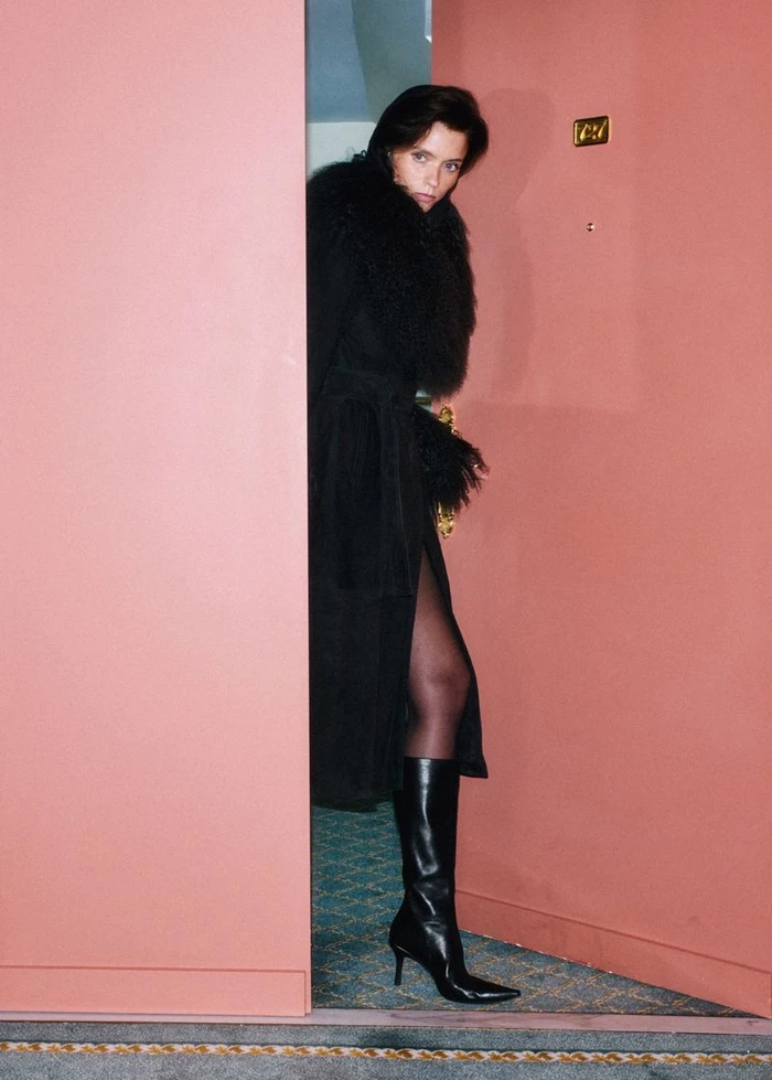 Model Abbey Lee in a black coat and boots opening a hotel door for Mango Fall 26
