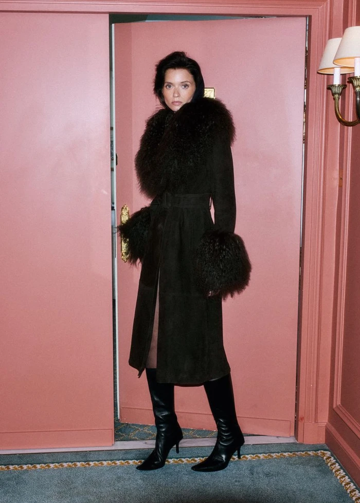 Abbey Lee wearing a dark fur-trimmed coat and black boots standing in a pink room for Mango