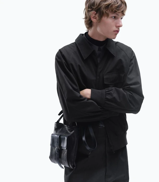 Model wearing a black jacket and carrying a black leather shoulder bag against a plain white background.