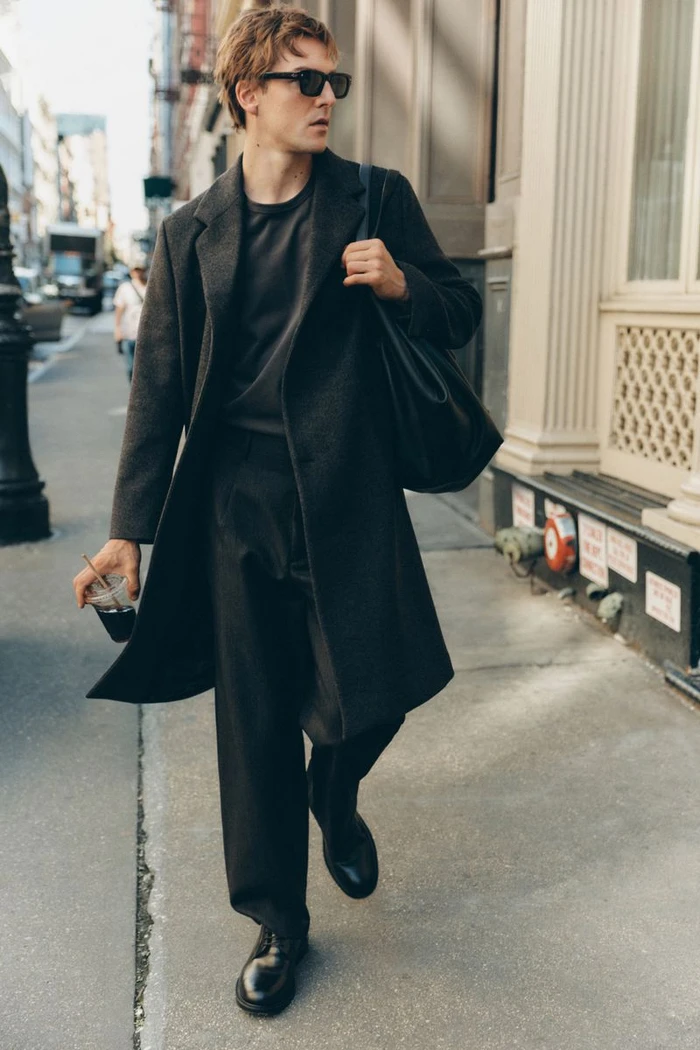 Man wearing dark sunglasses and a charcoal coat walking on a city sidewalk