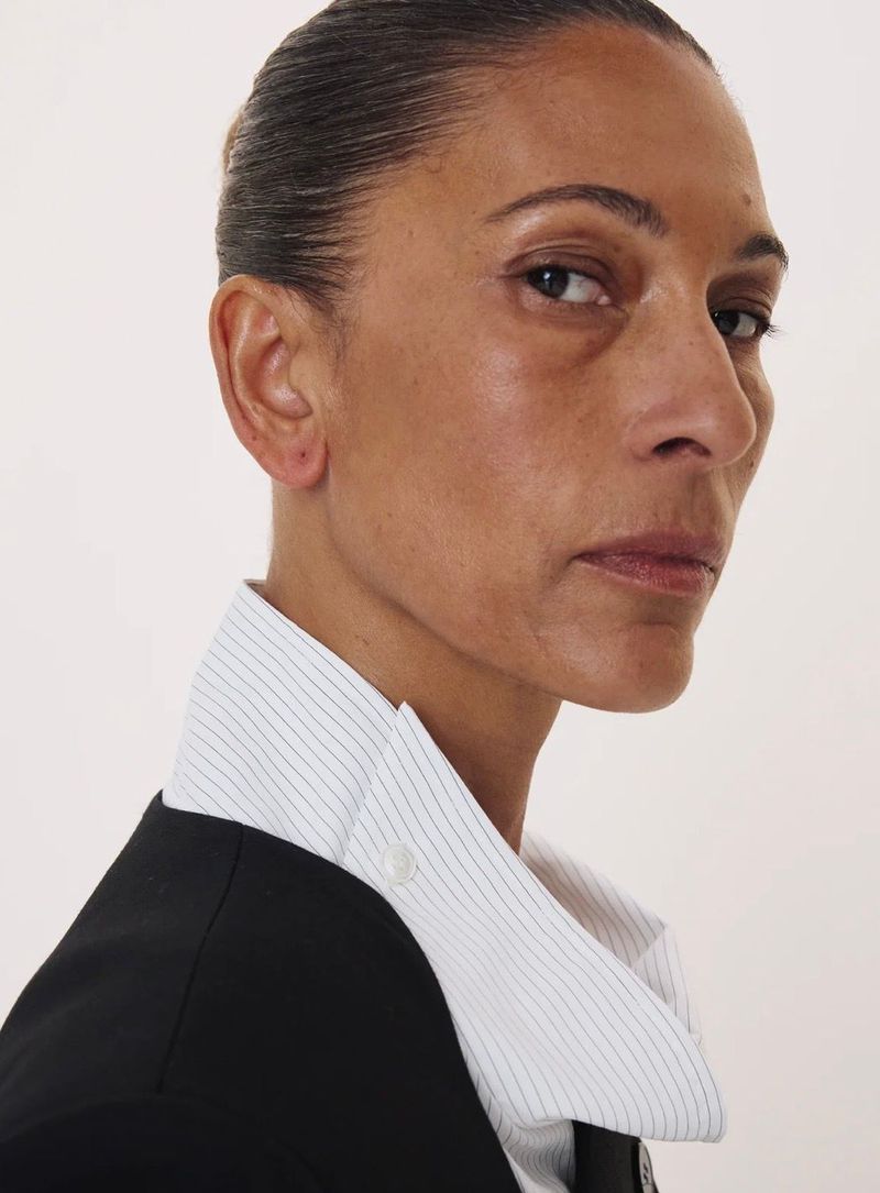 Close-up portrait of a woman in a black jacket and striped collared shirt against a white backdrop.
