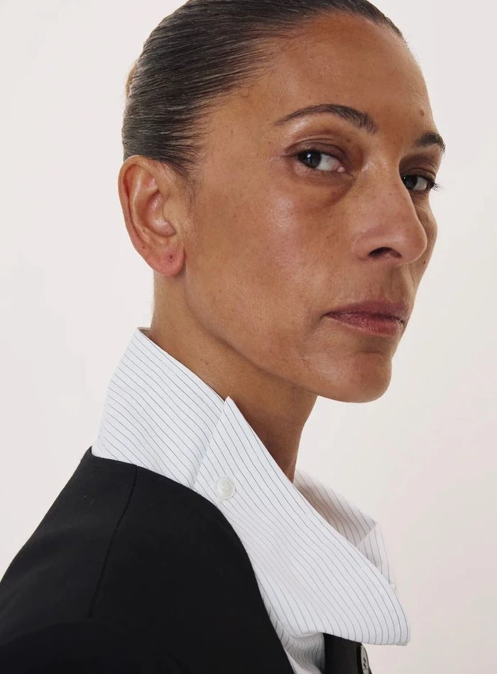 Close-up portrait of a woman in a black jacket and striped collared shirt against a white backdrop.
